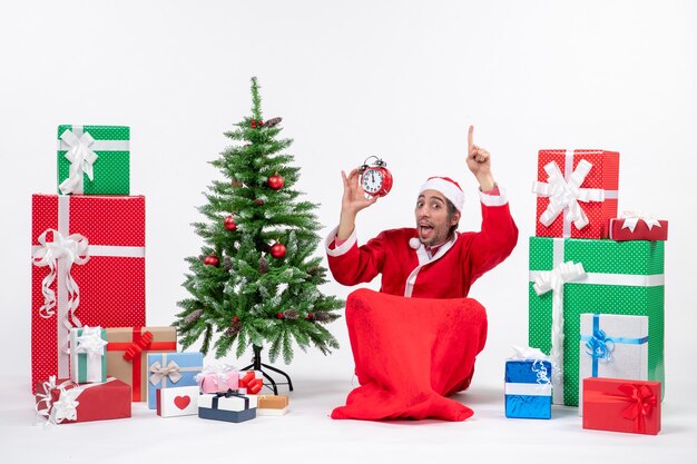 Эмоциональный сумасшедший удивленный санта-клаус сидит на земле и показывает часы, указывающие вверху возле подарков и украшенную рождественскую елку на белом фоне