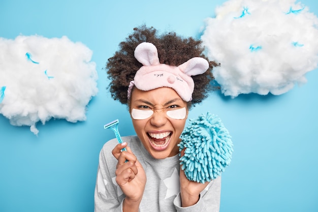 Emotional curly Afro American woman screams loudly wears sleepmask holds razor and sponge going to take shower dressed in nightwear poses against blue wall