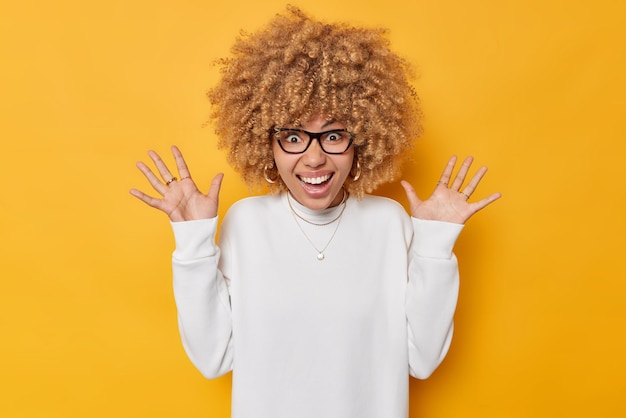 Emotional curly haired young European woman keeps palms raised feels very happy smiles toothily receieves awesome news wears white jumper and spectacles isolated over vivid yellow background