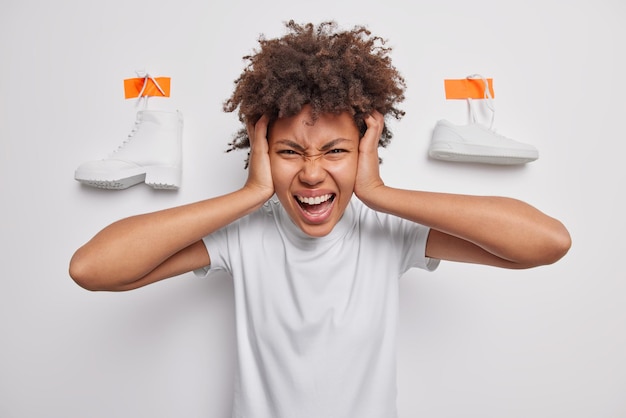 Free photo emotional curly haired young woman grabs head screams angrily expresses negative emotions wears casual t shirt stands against white background with plastered shoes being bothered by something