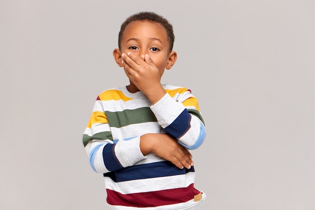 Эмоциональный милый афроамериканский мальчик, выражающий удивление или удивление, прикрывающий рот рукой в знак шока или скрытности, не шевелящий язык в голове. Истинные человеческие эмоции и реакция