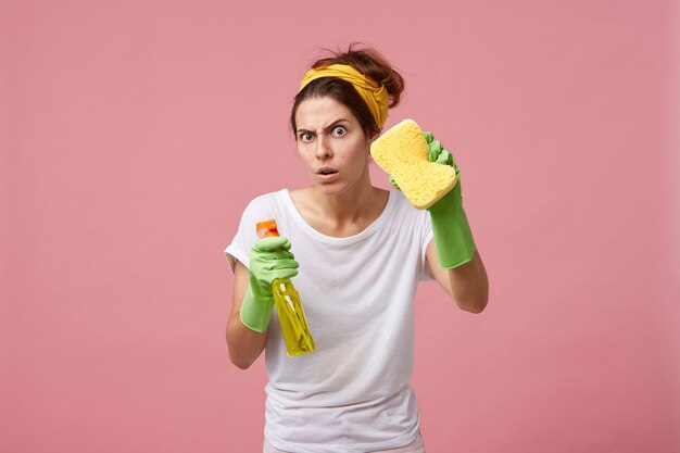 黄色のスポンジとスプレー洗剤を備えた緑のゴム手袋でポーズをとる感情的なかわいい女性は、彼女の顔に面白い表情で掃除をして家を掃除する準備ができています