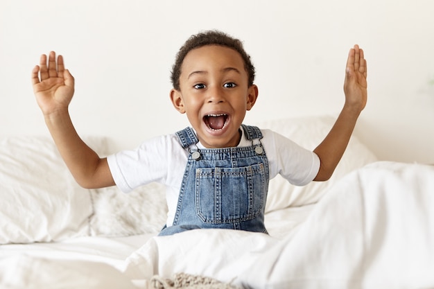 Free photo emotional cute little african american boy sitting on white bed