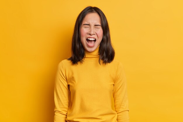 Emotional dark haired Asian woman screams loudly expresses rage keeps mouth widely opened wears casual yellow turtleneck in one tone with studio background. Human emotions and feelings concept