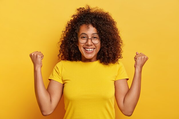 Emotional dark skinned woman makes hooray gesture, raises fists, smiles pleasantly, smiles amused, wears big round glasses and casual t shirt, has luminous curly hair, isolated over yellow background
