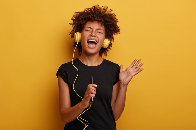 Emotional dark skinned woman with Afro hair raises arm, sings loudly, listens music which brings pleasant memories isolated on yellow