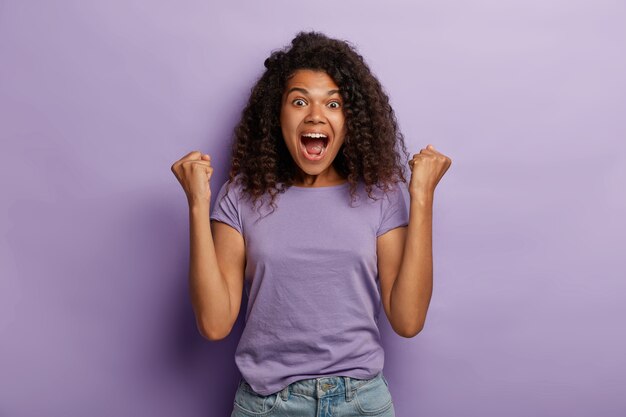 Emotional dark skinned woman with Afro hair, raises clenched fists, exclaims with excitemenet, rejoices sweet success, feels taste of victory, shouts for favourite team, wears purple t shirt and jeans