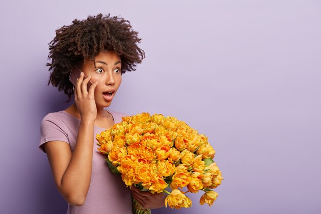 Emotional dark skinned young female has telephone conversation, impressed by shocking news, holds cellular near ear, gets aromatic yellow tulips, poses over purple wall, copy space on right side.