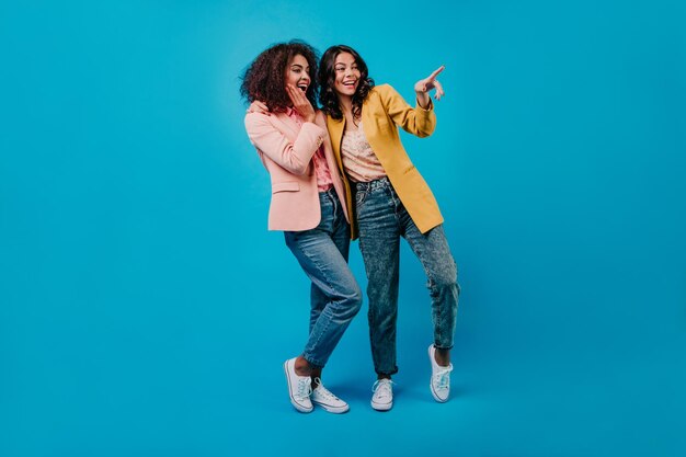 Emotional darkhaired ladies in jeans standing on blue background Indoor photo of two surprised girls