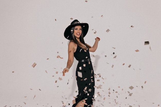 Emotional elegant woman wearing hat celebrating party in stylish black dress. Effective girl showing her lovely smile and dancing