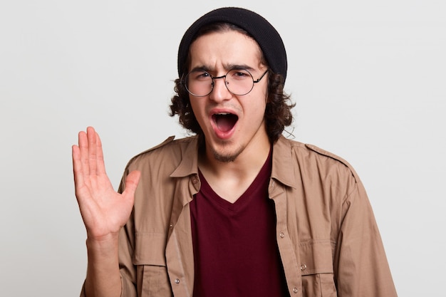 Emotional enthusiastic curly haired hipster raises one arm, opening his mouth widely with terror, having unpleasant facial expression. Screaming young guy wears dark red and beige shirts, black hat.