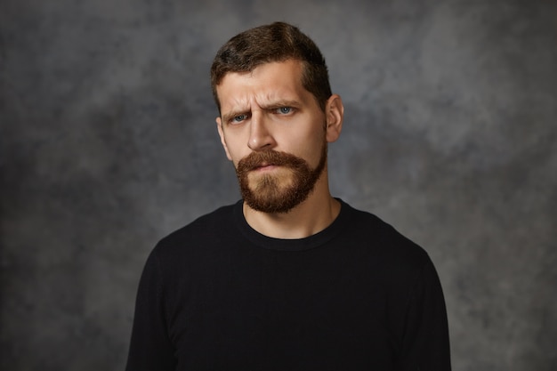  emotional European young male with fuzzy beard frowning, having displeased facial expression, being mad with his children for bad behavior, demonstrating annoyance and irritation