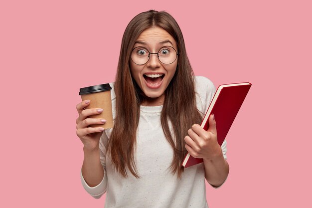 黒髪の感情的な興奮した若い女性は、幸せで叫び、持ち帰り用のコーヒーと本を運び、白いジャンパーを着て、ピンクの壁に孤立し、大喜びを感じます。感情の概念