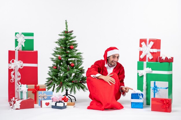 Emotional excited young man dressed as Santa claus with gifts and decorated Christmas tree sitting on the ground on white background