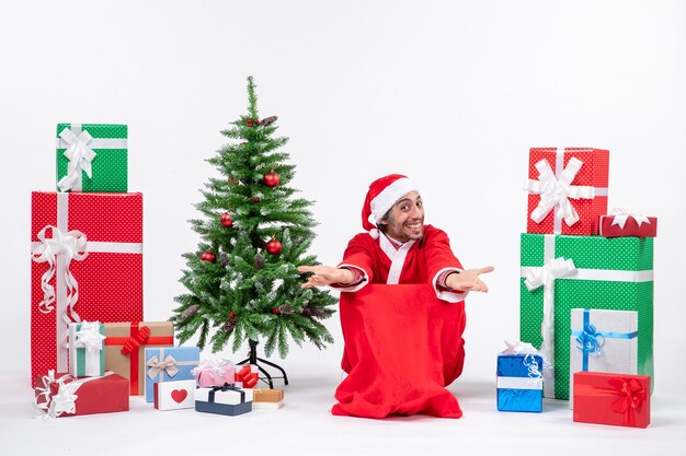 Emotional excited young man dressed as Santa claus with gifts and decorated Christmas tree welcoming someone on white background