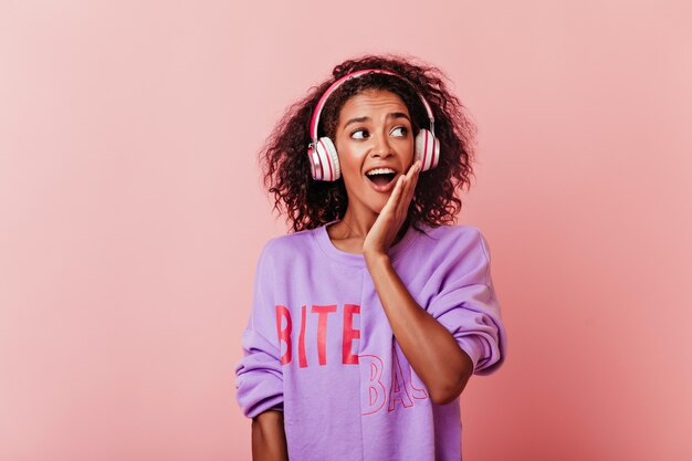 Emotional female model with black curly hair enjoying music. Happy african girl in headphones expressing amazement.