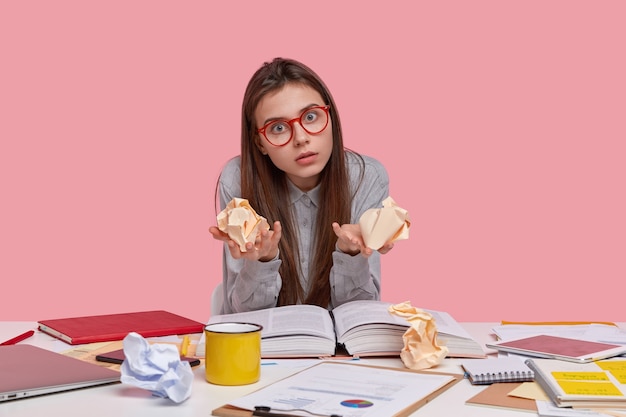 Emotional female nerd has shocked gaze directly at camera, stares through spectacles, holds crumpled paper in both hands, writes financial report