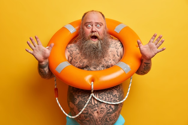 Emotional frightened bearded man raises hands and screams loudly, has bugged eyes and opened mouth, has tattooed body, stands with inflated lifebuoy, afraids of swimming, poses indoor alone.