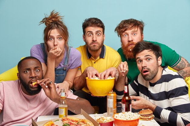 Emotional frightened millennial friends spend free time together, watch online thrilling movie, eat tasty pizza, drink beer, have interest gaze, isolated against blue wall, enjoy television.