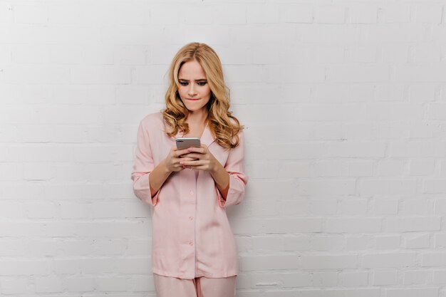 Эмоциональная девушка в хлопковой пижаме позирует с телефоном на белой стене. Прекрасная кудрявая женщина в розовом ночном костюме, глядя на экран смартфона.