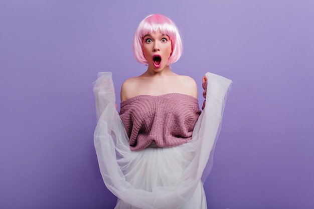 Free photo emotional girl in shiny peruke plays with her white skirt. indoor portrait of surprised young woman with pink hair isolated on purple wall.