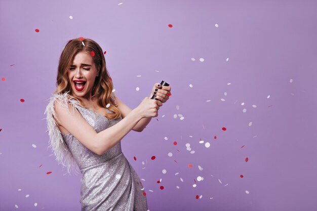 Emotional girl with red lips dressed in silver outfit throwing confetti Excited woman in shiny dress smiling on purple isolated backdrop