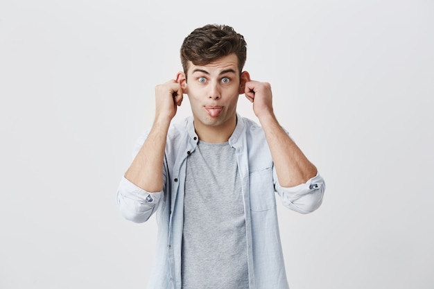 Emotional gloomy young male student in blue shirt over gary t-shirt grimacing, mocking, making faces, pulling his ears, sticking outhis tongue, looking  with stupid expression on his face.