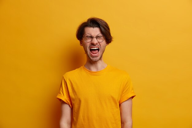 Emotional guy keeps mouth wide opened, screams from disappointment, shouts with closed eyes, feels troubled as lost huge bet, dressed in bright yellow t shirt, looses temper, has life problems