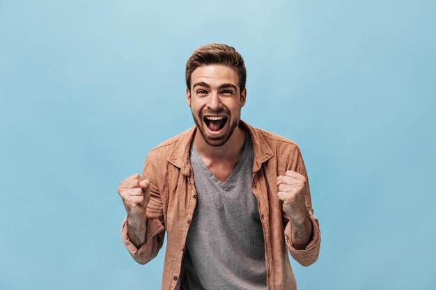 Emotional guy with beard in brown shirt and grey plain tshirt rejoicing and looking into camera on isolated blue background