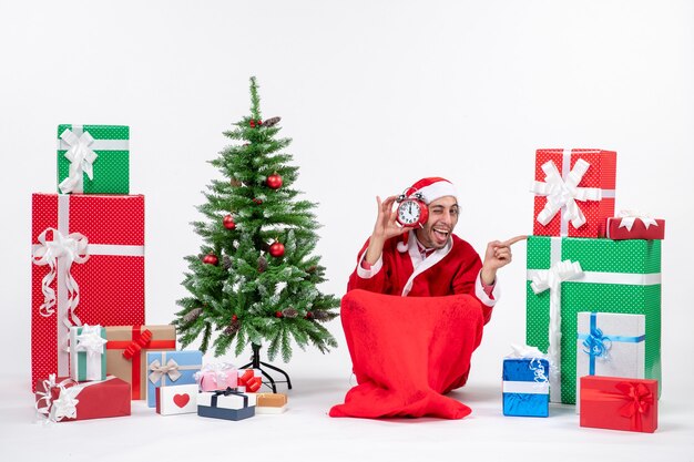 Emotional happy funny santa claus lsitting on the ground and showing clock near gifts and decorated xsmas tree on white background