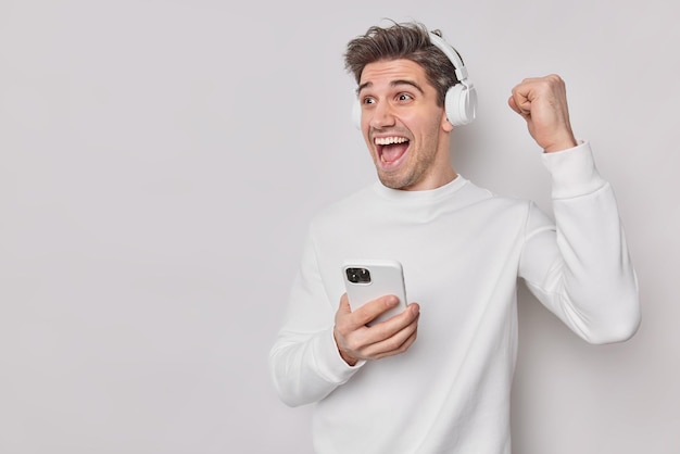 Emotional happy man rejoices awesome news clenches fist celebrates achievement dressed in casual jumper listens favorite audio track isolated over white background. People fun and leisure concept