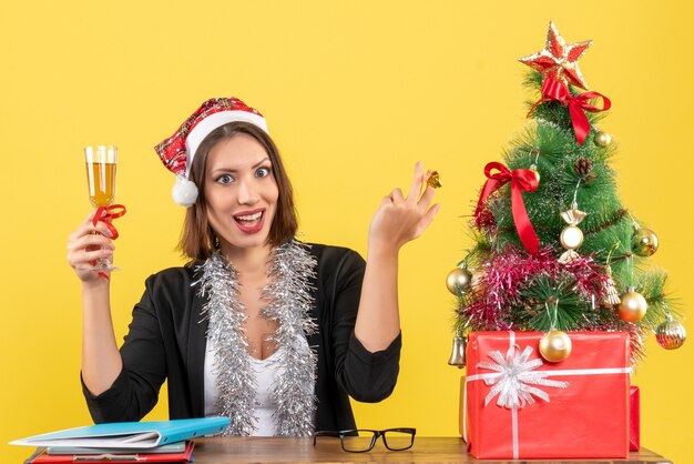 Emotional happy satisfied charming lady in suit with santa claus hat and new year decorations raising wine in the office on yellow isolated 