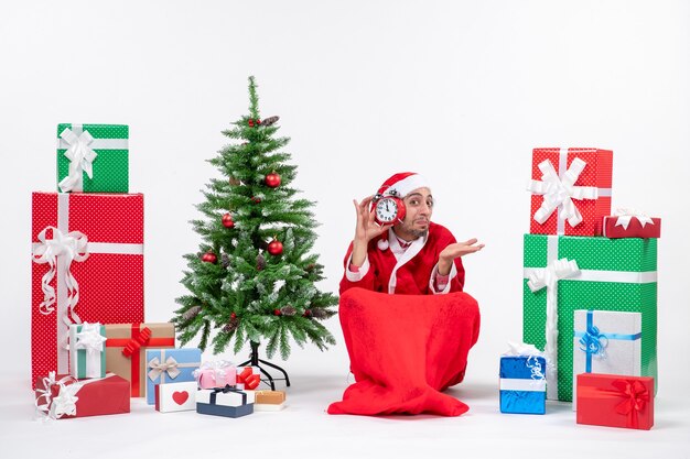 Emotional happy surprised santa claus sitting on the ground and showing clock near gifts and decorated xsmas tree on white background