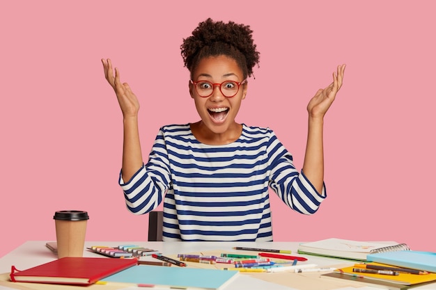 Emotional illustrator raises hands in eureka, exclaims with happiness, has nice idea for masterpiece, works at table with notepad, crayons, coffee, wears striped sweater, isolated over pink wall