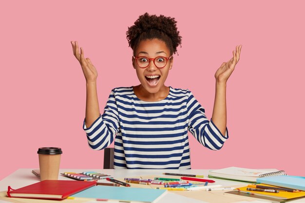 감성적 인 일러스트 레이터가 유레카에서 손을 들고 행복하게 외치고 걸작에 대한 좋은 아이디어를 가지고 있으며 메모장, 크레용, 커피가있는 테이블에서 작업하고, 줄무늬 스웨터를 입고, 분홍색 벽 위에 절연