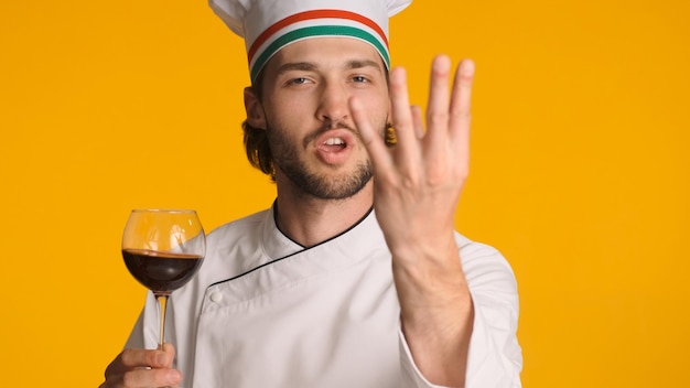 Emotional Italian chef with glass of red wine showing delicious gesture over colorful background Man sommelier tasting good wine