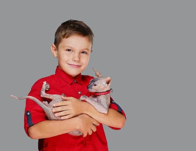 빨간 폴로 티셔츠를 입은 감정적인 소년은 고양이를 안고 있습니다. 회색 배경에 고립.