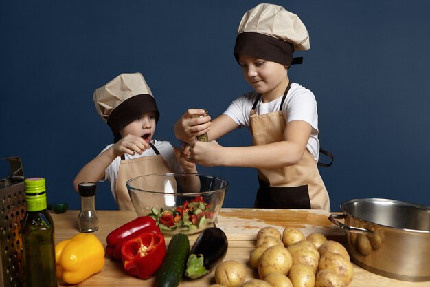 Emotional little boy standing at kitchen table with potatoes, peppers, cucumber and eggplant, opening mouth excitedly, watching his elderly brother putting too much spice into bowl of salad