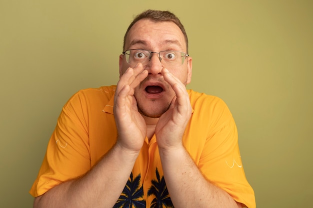 Emotional man in glasses wearing orange shirt shouting with hands near mouth standing over light wall