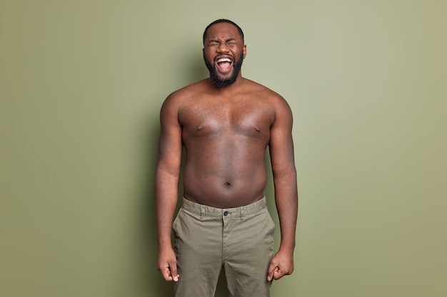 Free photo emotional man with naked torso wears shorts screams loudly opens mouth widely has thick beard poses against khaki color wall keeps arms down