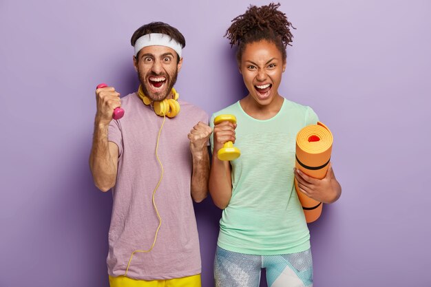 Emotional mixed race woman and man shout loudly, hold karemat and weights, have training with coach, yell from despair, being tired of workout, isolated on purple wall. People, sport, lifestyle