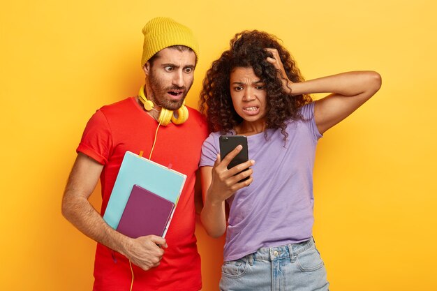 Emotional nervous mixed race students read shocking information from website, stare at smartphone, carry notepad