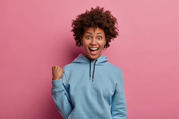 Emotional overjoyed curly woman raises clenched fist, has upbeat mood, shouts for favourite football team, wears casual blue hoodie, gets excellent news, reacts on something awesome, stands indoor