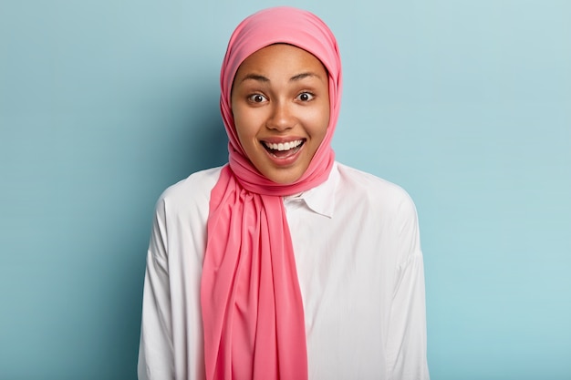 Эмоциональная восторженная мусульманка счастливо смеется, выражает радость, выражает искренние чувства, носит розовый головной убор, показывает белые зубы, изолированные на синей стене. Концепция человеческих эмоций