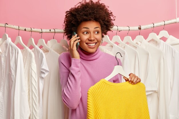 感情的な大喜びの女性は電話をかけ、白い服でいっぱいのワードベラックの近くに立ち、ニットの冬の黄色いセーターを持って、ファッションモールで買い物を楽しんでいます。