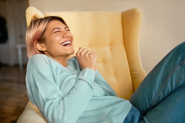 Emotional overjoyed young European woman in jeans and blue sweatshirt lying on armchair keeping eyes closed