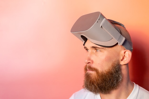 Emotional portrait of a man with a beard wearing virtual reality glasses in the studio on a pink orange background