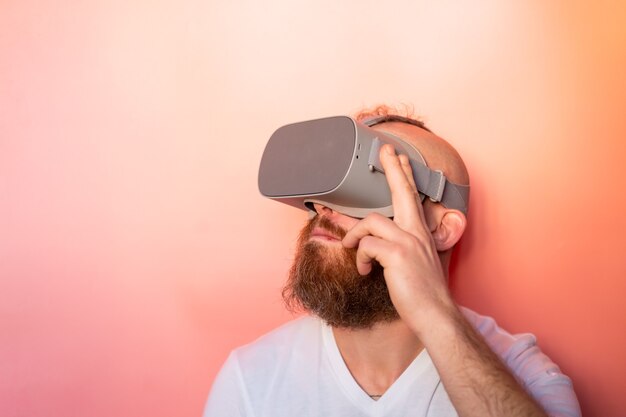 Emotional portrait of a man with a beard wearing virtual reality glasses in the studio on a pink orange background