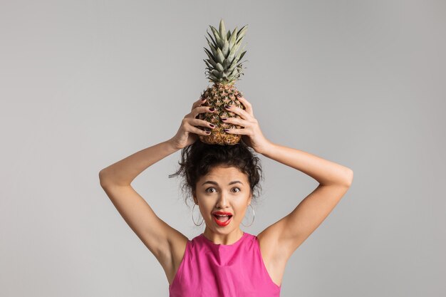 Free photo emotional portrait of young exotic brunette woman in pink shirt holding pineapple on her head