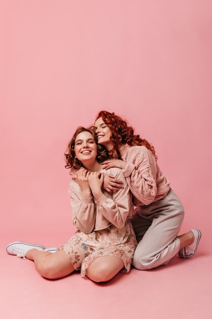 Emotional pretty girls sitting on pink background. Two friends embracing while posing on floor.